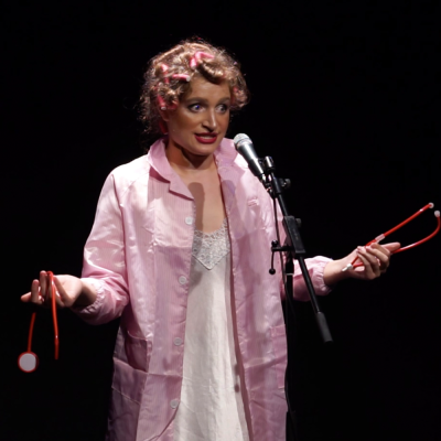 A young woman in a pink lab coat with her hair in curlers holds a broken stethoscope